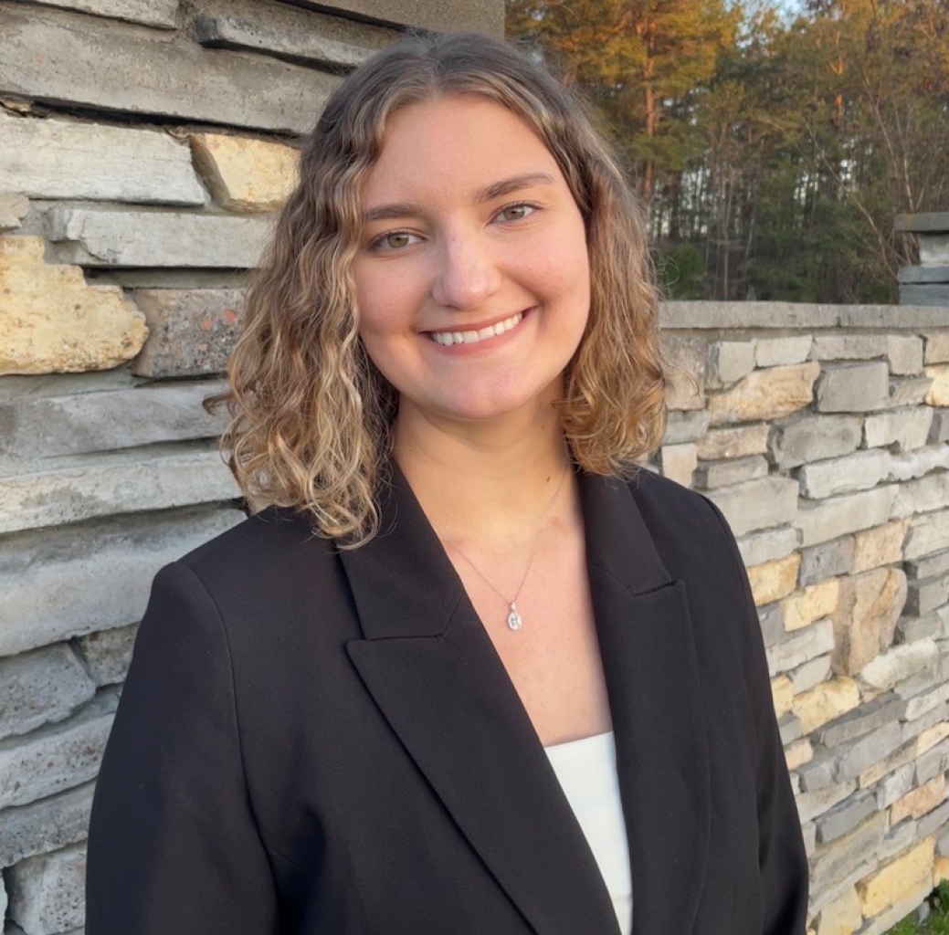 Rosemary Faircloth wearing a black blazer over a white shirt in front of a wall of stone bricks with trees in the background.